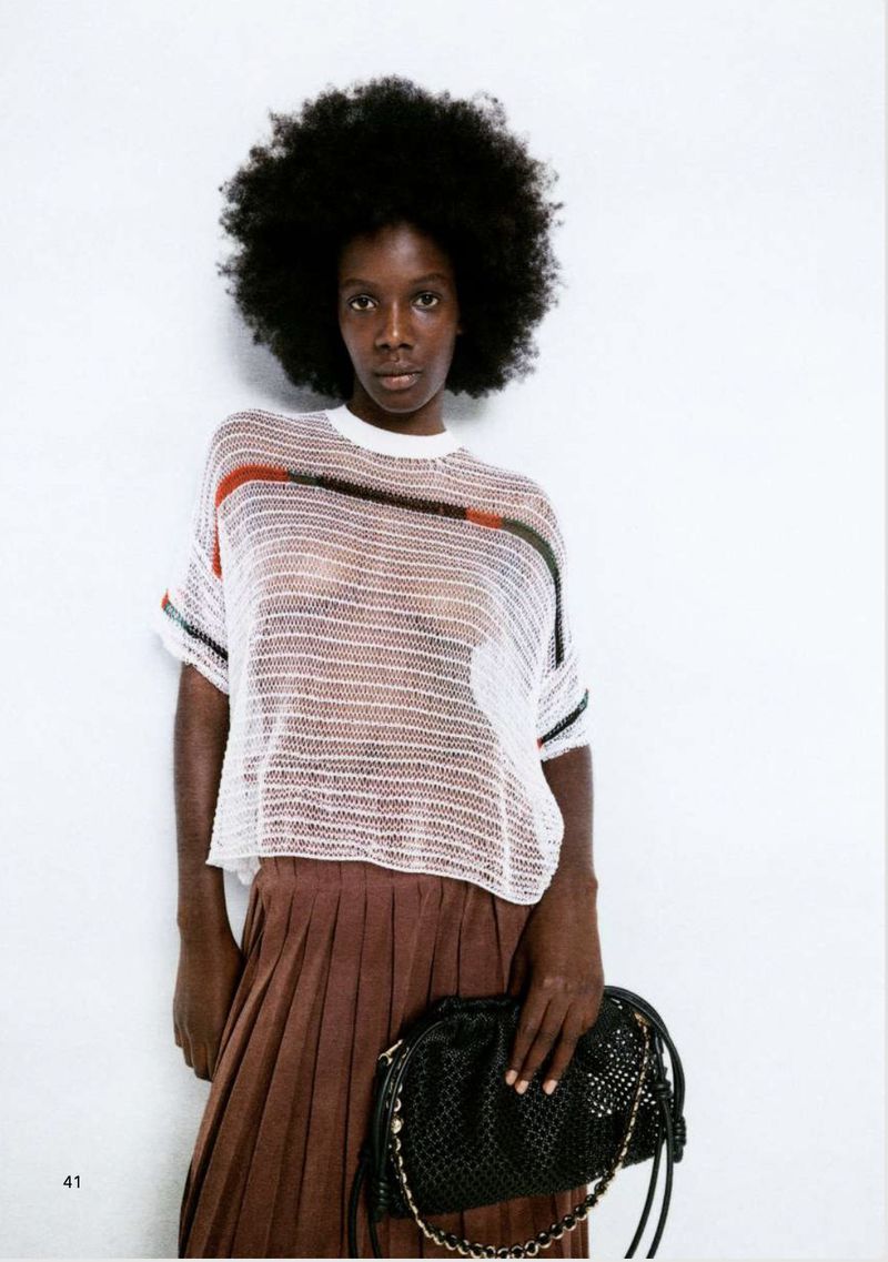 Fashion editorial shot of a Black woman with an afro, wearing a white knitted shirt, brown skirt and holding a black bag against a white background.