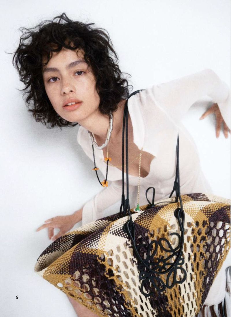 Fashion editorial shot of a young woman with curly hair wearing a white top, necklaces, and holding a patterned woven bag.