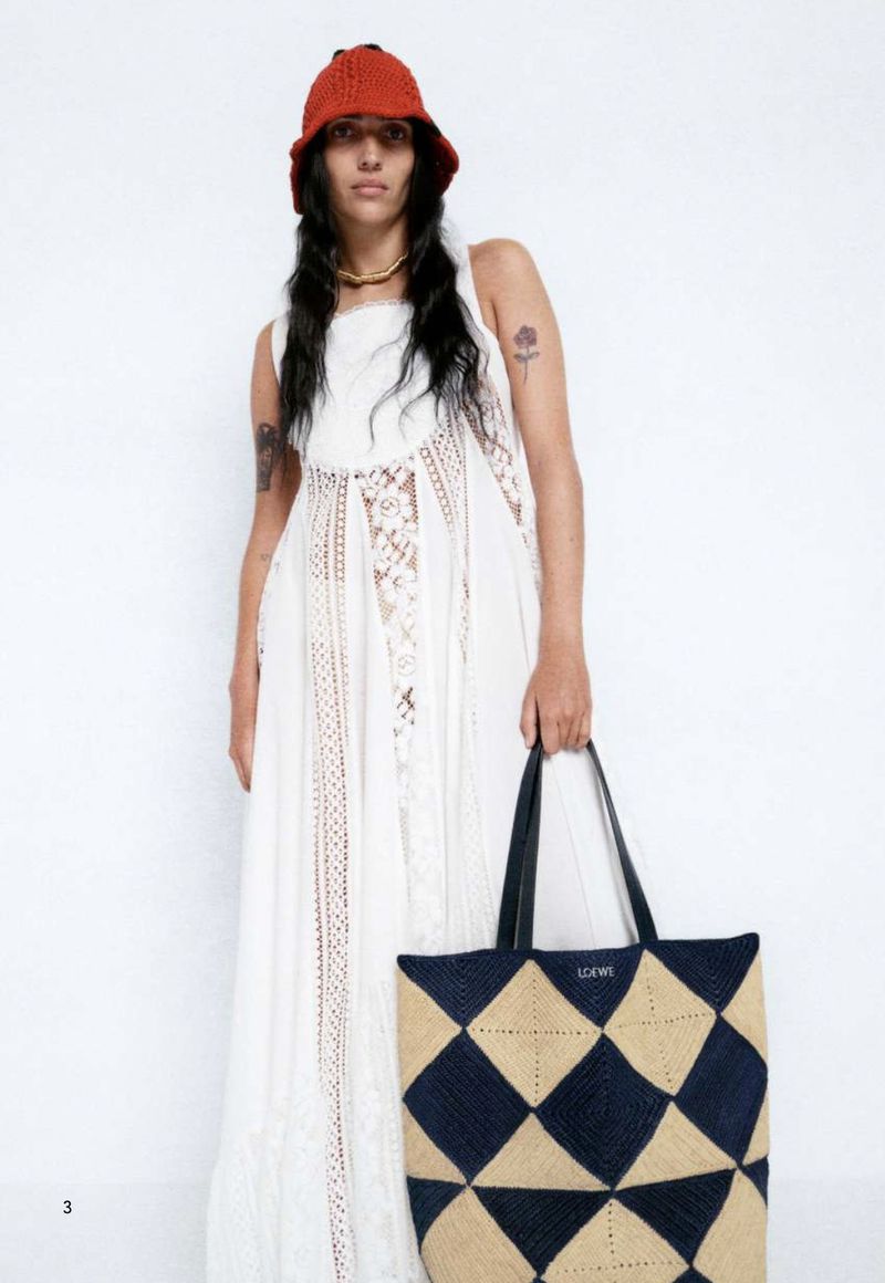 A woman in a white dress holds a large tote bag with a diamond pattern. She wears an orange bucket hat and a gold necklace.