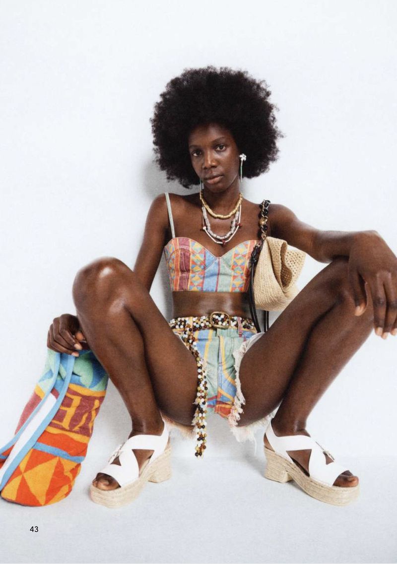 A woman with an afro hairstyle poses while sitting down, wearing a patterned crop top and shorts, along with platform sandals.