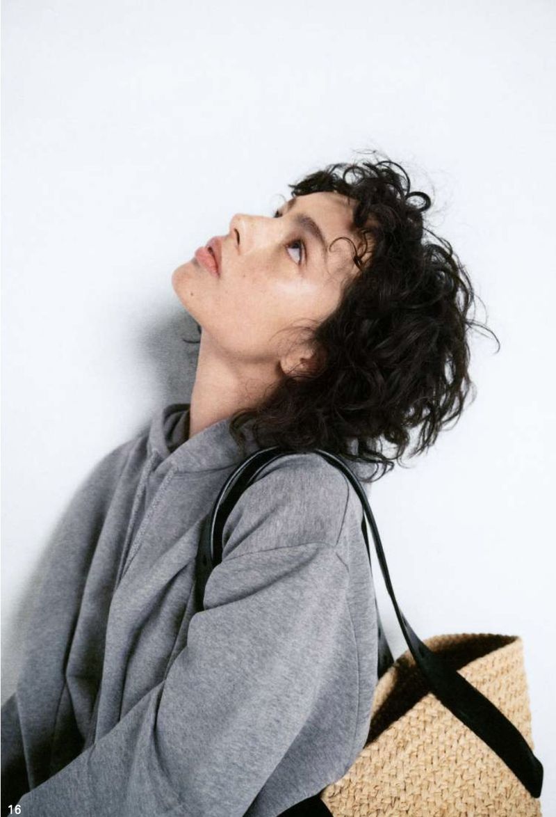 Young woman in grey hoodie looks up. Her short curly hair and straw bag with black straps add a touch of casual chic against a white background.
