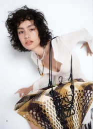 Model with dark curly hair wearing a white top and holding a large patterned woven straw bag against a white background
