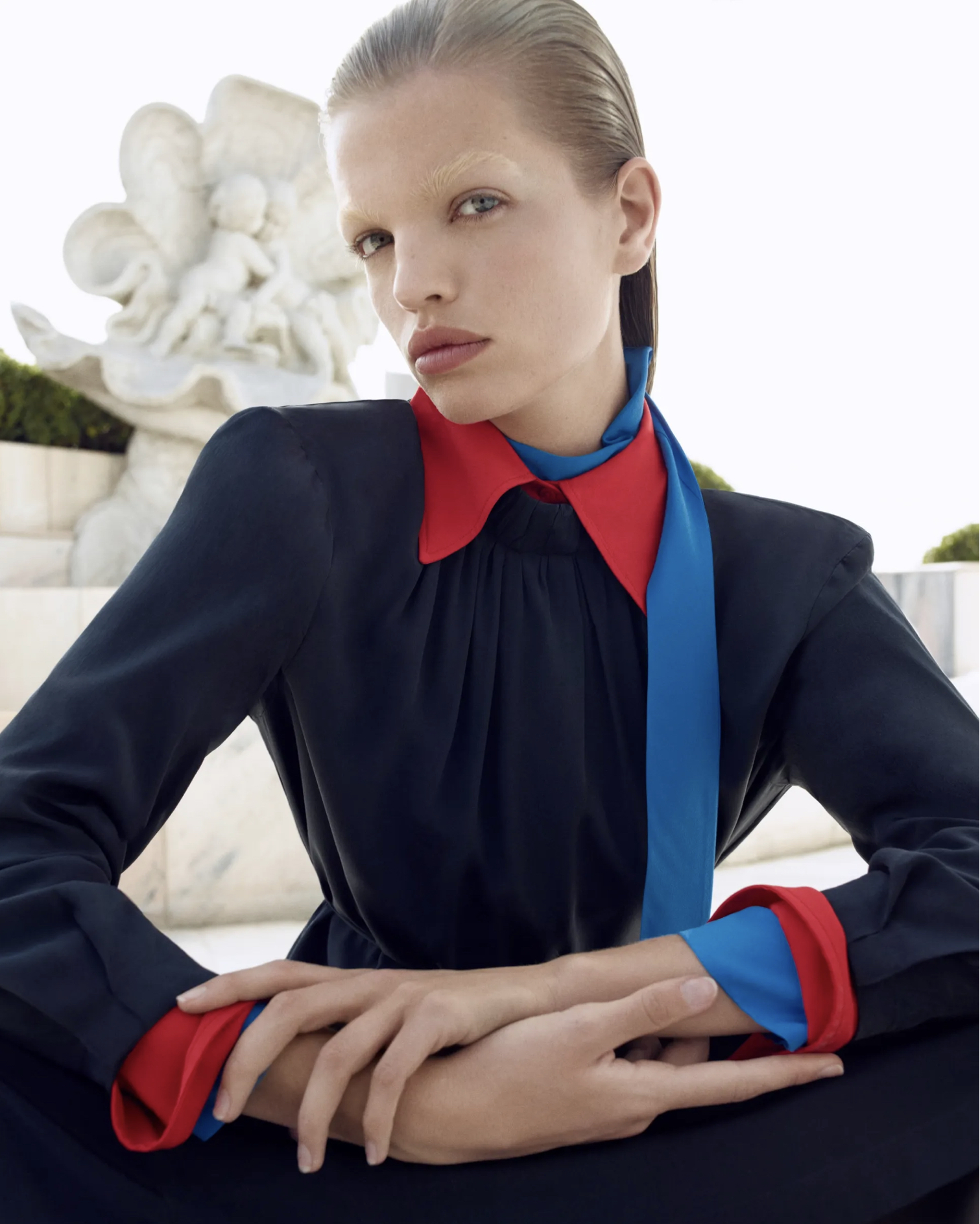 Portrait of a fashion model in a navy dress with red and blue accents, posing outdoors near a white sculpture on a bright day.