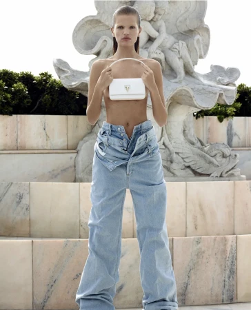 Fashion shot of a female model holding a white purse, posing on marble steps with a sculpted marble fountain in the background.