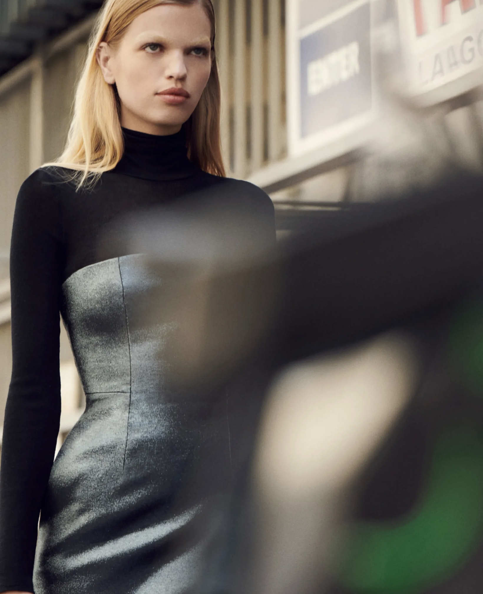 A fair-skinned woman with blonde hair poses in a fitted metallic-silver dress with a black top in an urban setting.