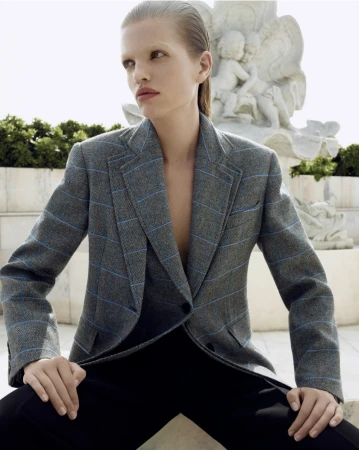 Blonde fashion model poses in a gray blazer and black pants in front of a marble cherub statue in an outdoor setting. 
