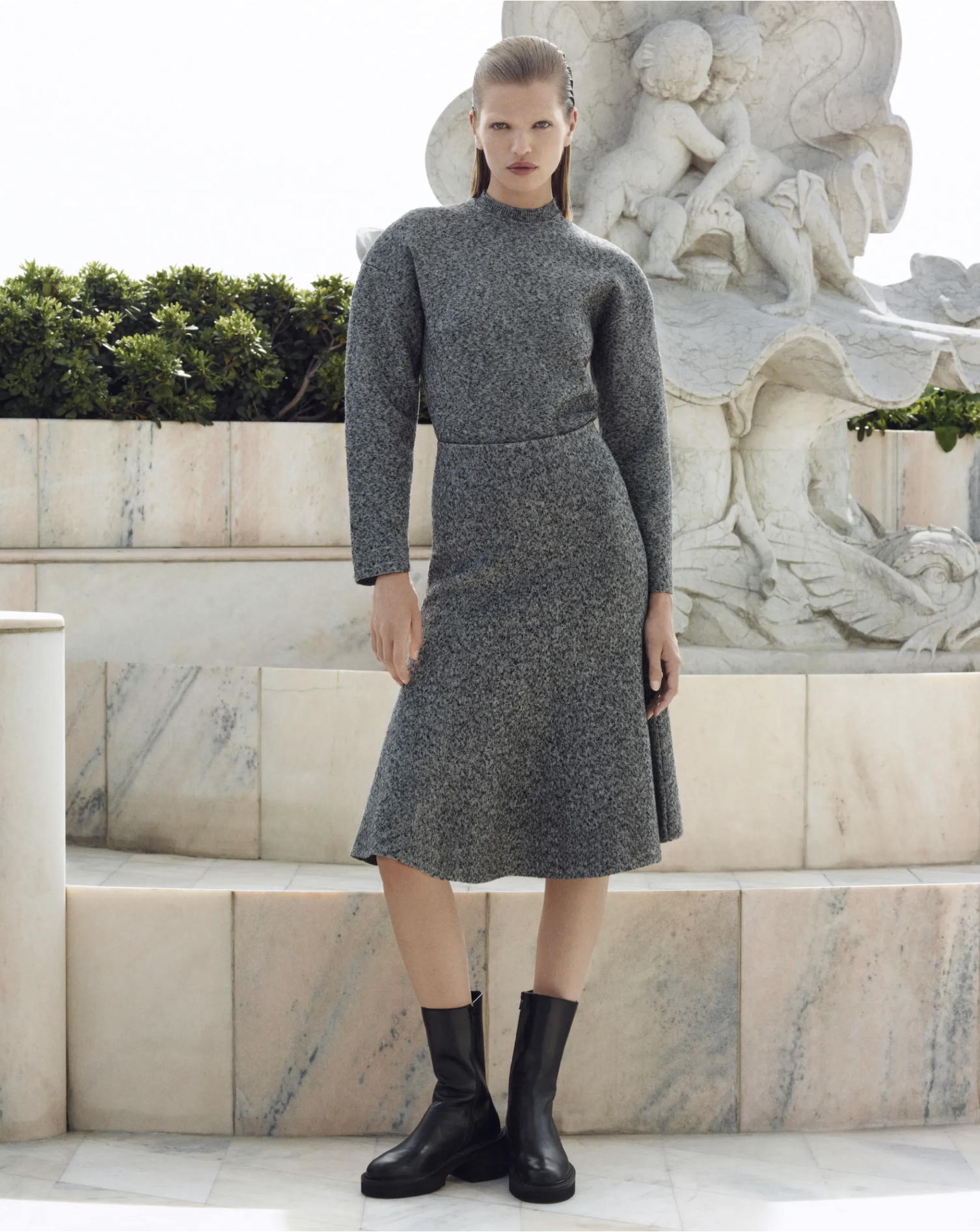 Fashion model poses outdoors in gray knit dress and black boots near a marble sculpture, showcasing a modern yet classic style.