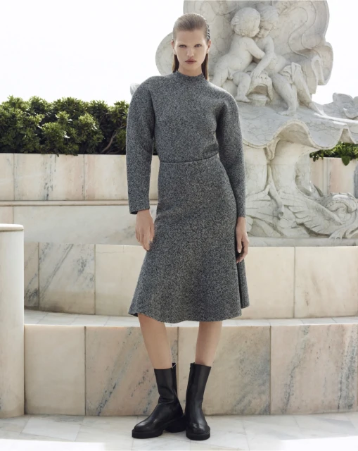 Fashion model poses outdoors in gray knit dress and black boots near a marble sculpture, showcasing a modern yet classic style.