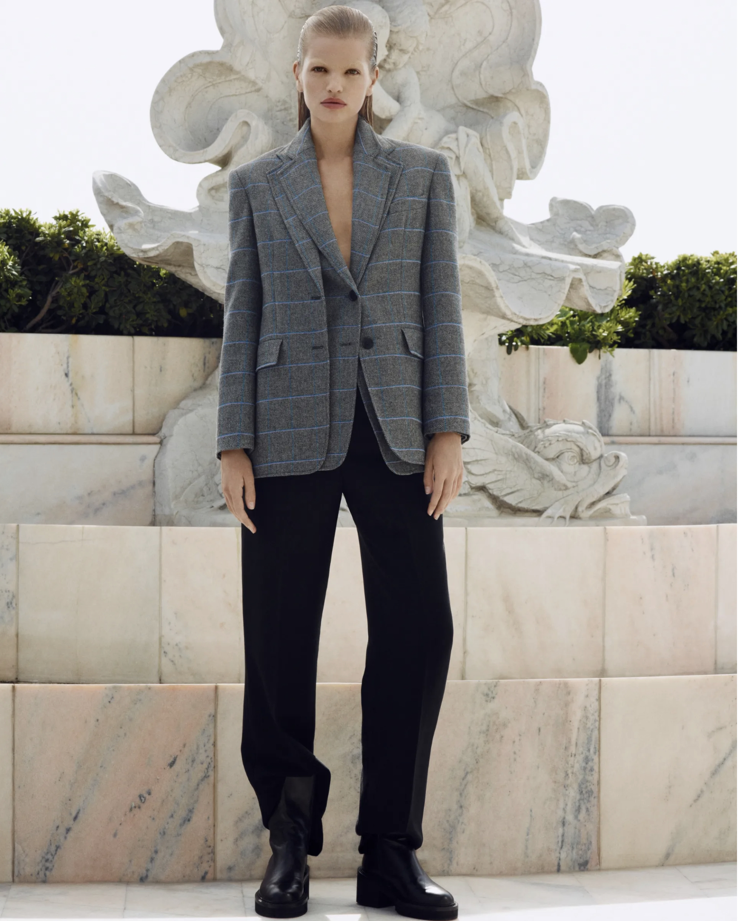 Fashion editorial shot: Woman poses in an oversized grey blazer, black pants, and chunky boots in front of a marble fountain.