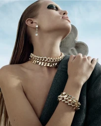 Model Daphne Groeneveld wearing layered gold chain necklaces and a grey tweed blazer against a blue sky