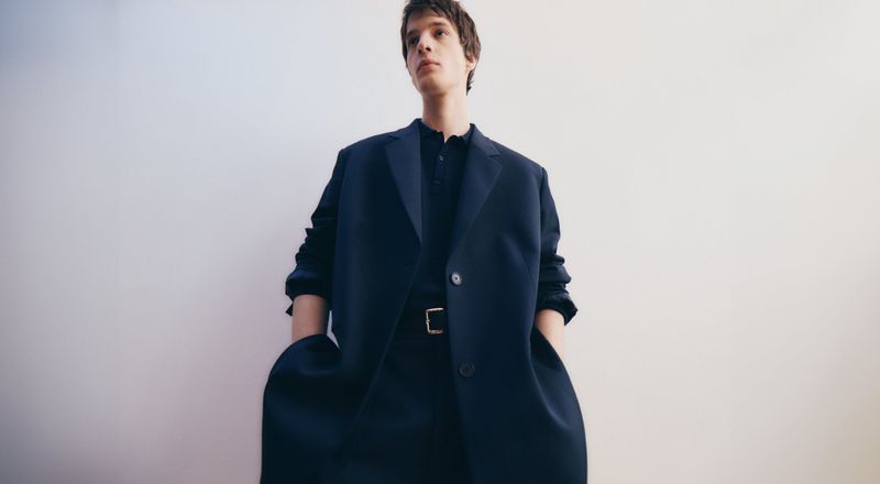 Man in dark blue oversized coat and matching suit poses against a light background in a fashion editorial, looking up and away.