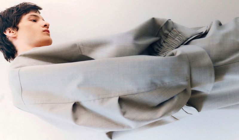 Fashion shot of a young adult man in a light grey jacket lying down, angled towards the camera against a solid white background.