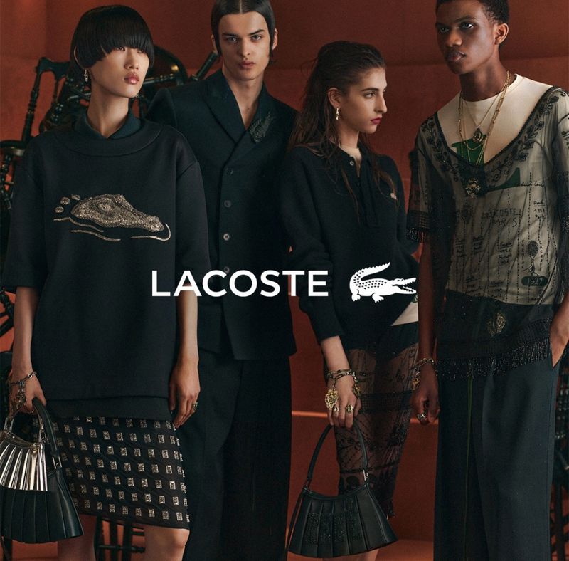 Lacoste fashion ad featuring four models in dark clothing and pleated accessories, against a blurred orange backdrop with metal details.