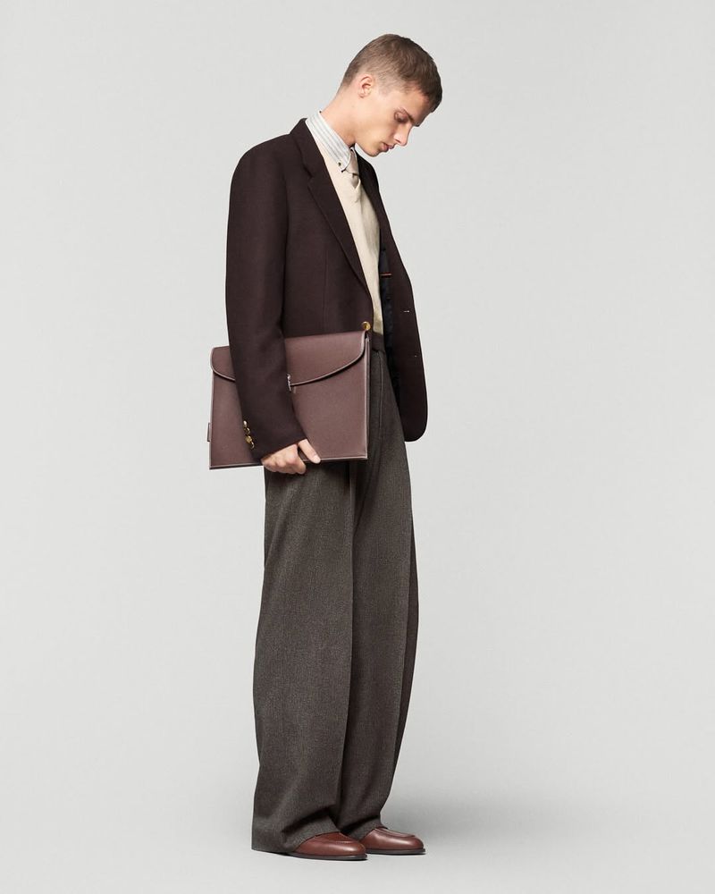 A slender adult male poses in a dark brown blazer, gray wool trousers, and brown leather shoes, holding a brown leather briefcase against a light gray studio backdrop.