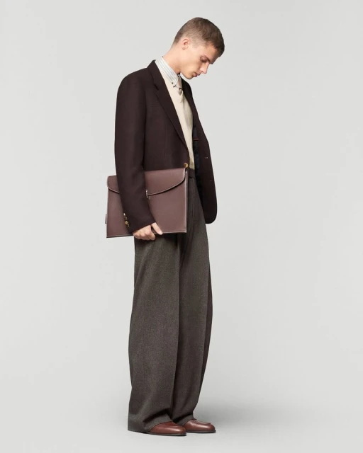 A slender adult male poses in a dark brown blazer, gray wool trousers, and brown leather shoes, holding a brown leather briefcase against a light gray studio backdrop.