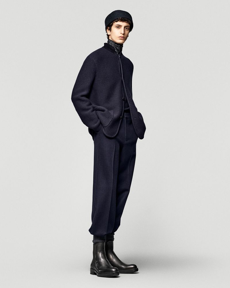 Male model poses in a dark blue jacket, trousers, and black boots in a studio setting with a plain backdrop. Fashion editorial shot.