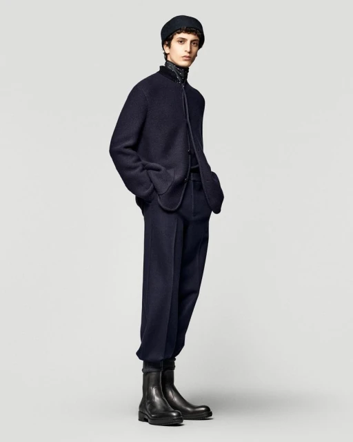 Male model poses in a dark blue jacket, trousers, and black boots in a studio setting with a plain backdrop. Fashion editorial shot.