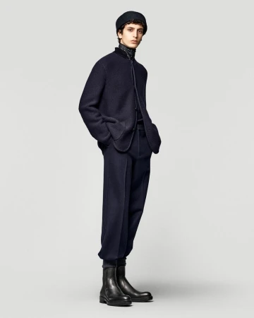 Male model poses in a dark blue jacket, trousers, and black boots in a studio setting with a plain backdrop. Fashion editorial shot.