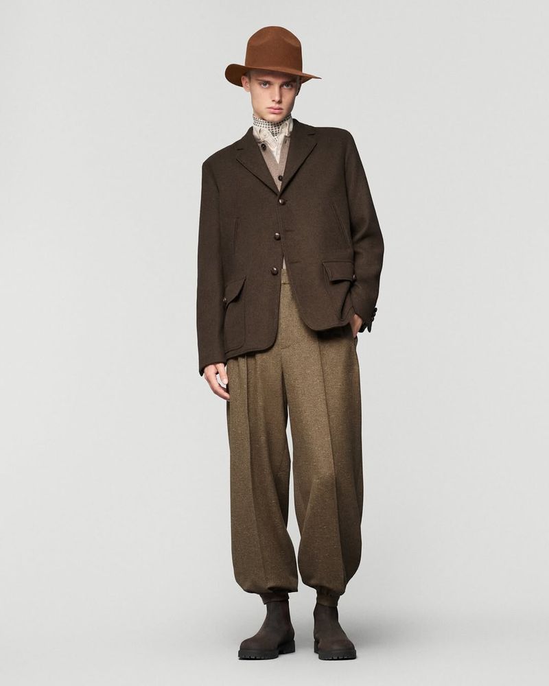 Full-length studio shot of an adult male wearing a brown hat, jacket, and pants. The man poses against a neutral grey background.