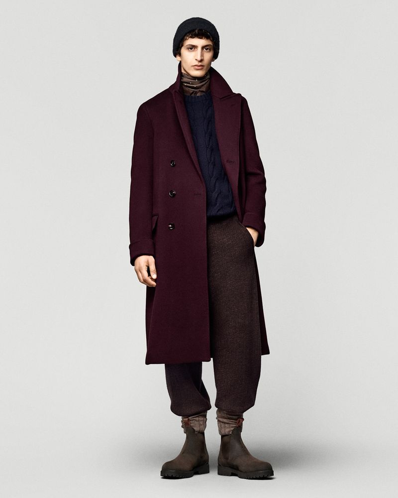 Full-length shot of adult male fashion model posing in a burgundy overcoat, navy sweater, brown pants, black knit hat, and leather boots.