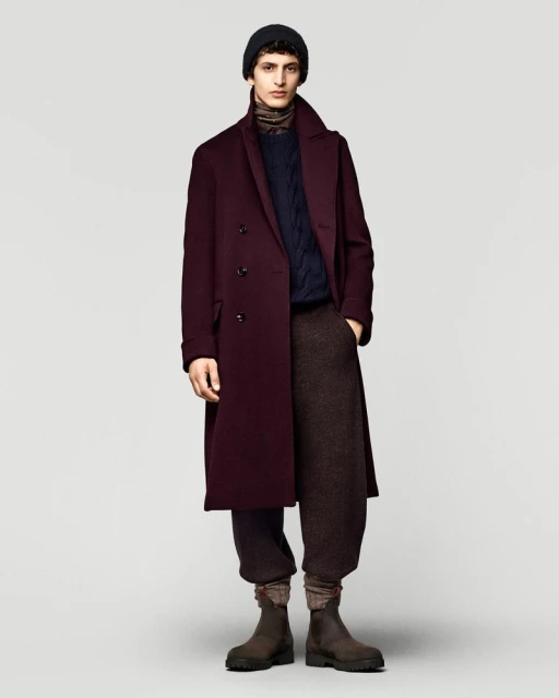 Full-length shot of adult male fashion model posing in a burgundy overcoat, navy sweater, brown pants, black knit hat, and leather boots.
