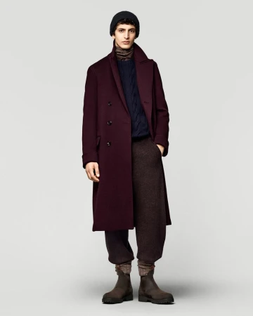 Full-length shot of adult male fashion model posing in a burgundy overcoat, navy sweater, brown pants, black knit hat, and leather boots.