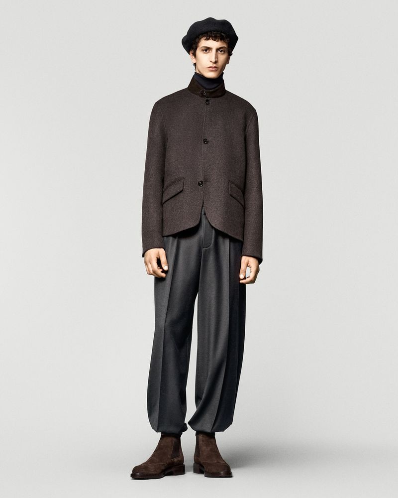 Male fashion model in a brown jacket, black turtleneck, grey trousers, and brown boots, posing in a studio with a light grey background.