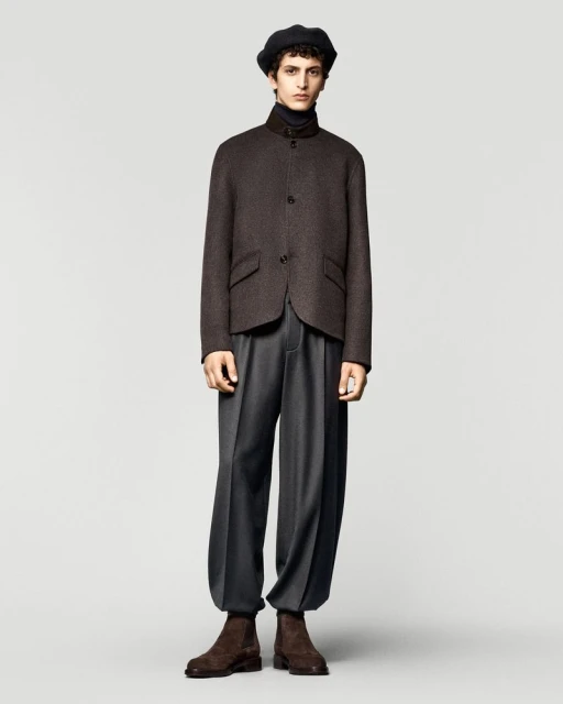 Male fashion model in a brown jacket, black turtleneck, grey trousers, and brown boots, posing in a studio with a light grey background.