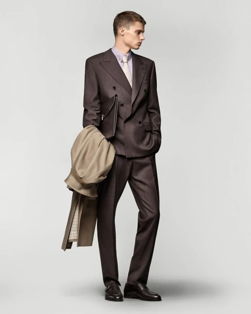 Full-length shot of a male model in a dark brown suit with light shirt and tie, holding a beige overcoat in a neutral indoor setting.