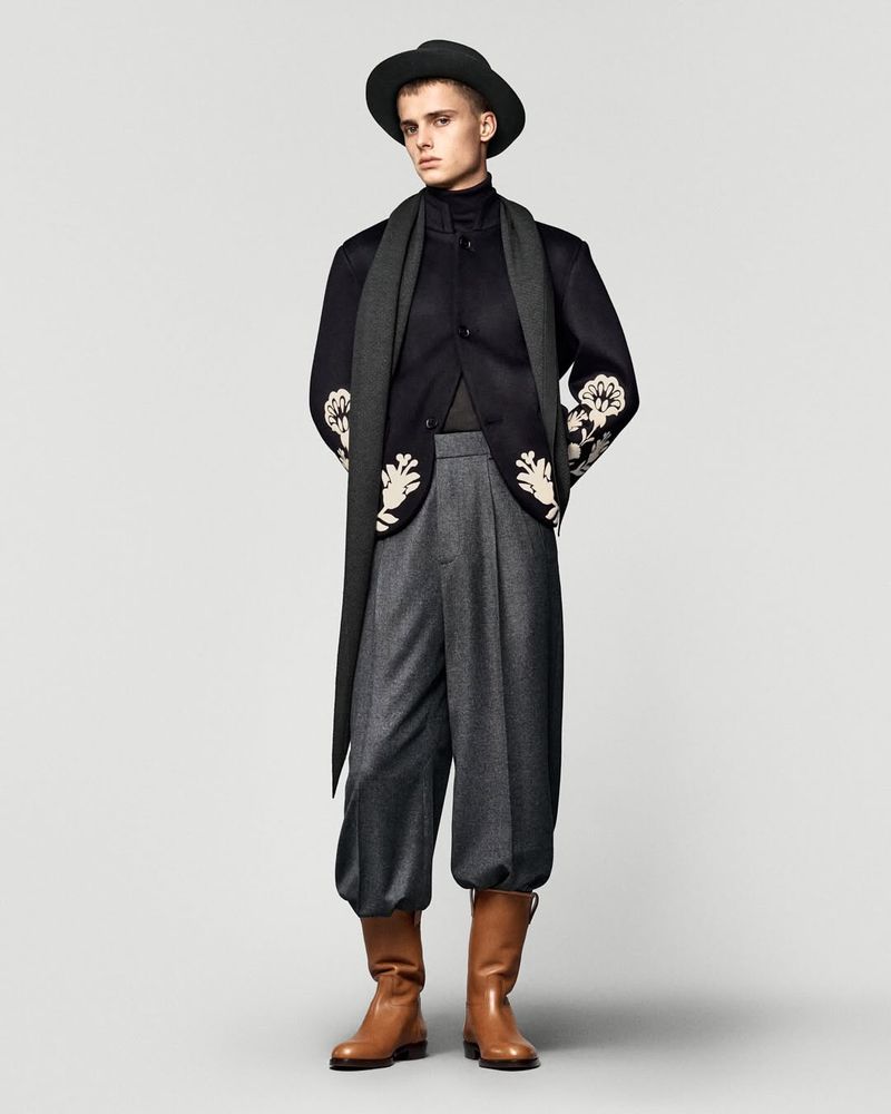 Full-length studio shot of a young adult male in a hat, scarf, jacket, trousers, and boots, against a neutral backdrop.