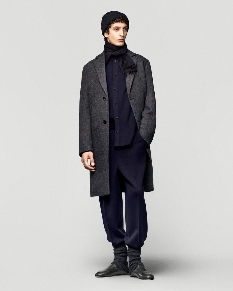 Adult male fashion model in a studio shot wearing a dark grey wool overcoat, knit cap, scarf, dress shirt, and jogger-style pants with leather dress shoes.