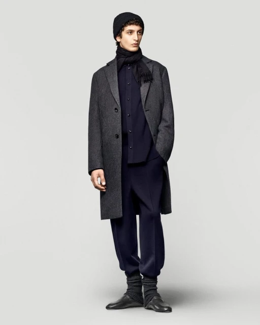 Adult male fashion model in a studio shot wearing a dark grey wool overcoat, knit cap, scarf, dress shirt, and jogger-style pants with leather dress shoes.