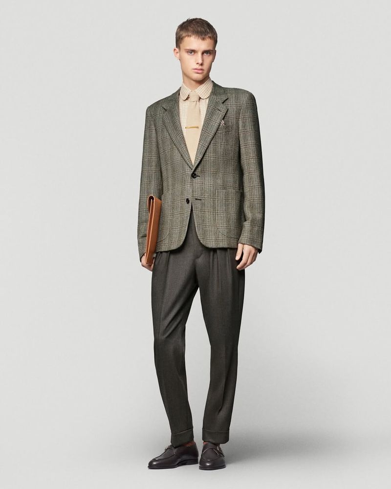 A young adult male stands in a studio, wearing a tweed blazer, tan collared shirt, grey trousers, and leather loafers, holding a briefcase.