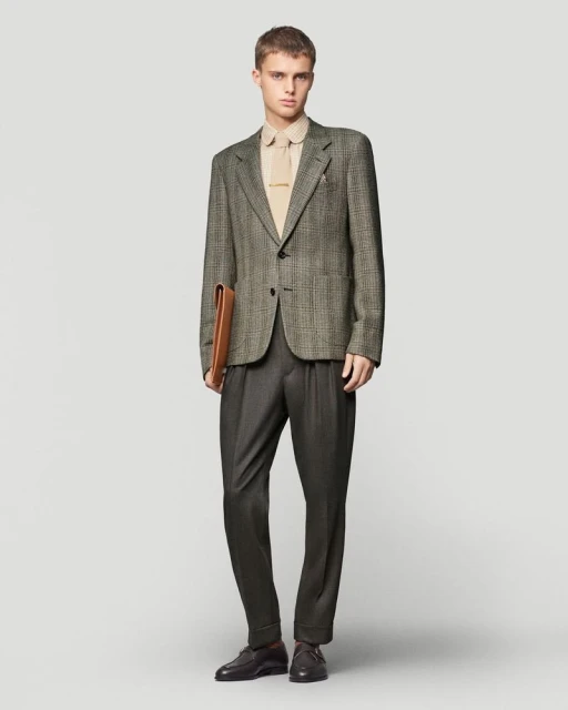 A young adult male stands in a studio, wearing a tweed blazer, tan collared shirt, grey trousers, and leather loafers, holding a briefcase.