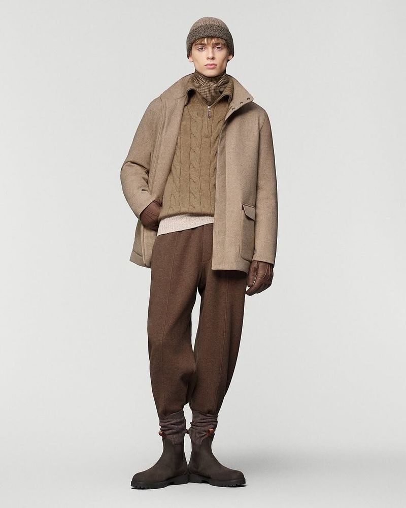 Full-length studio shot of a male fashion model wearing a beige jacket, cable-knit sweater, brown pants, hat, gloves, and boots on a white background.