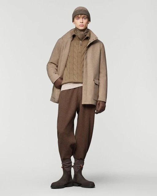 Full-length studio shot of a male fashion model wearing a beige jacket, cable-knit sweater, brown pants, hat, gloves, and boots on a white background.
