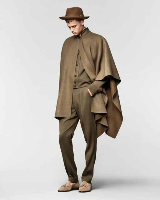 Male fashion model poses in neutral studio wearing a brown cape, suit, and fedora hat, showcasing a blend of modern and classic style.