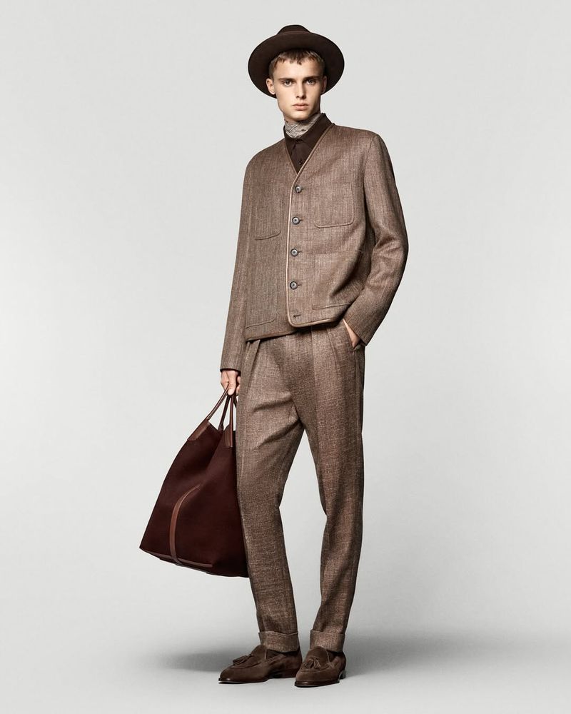 Fashionable adult male in a tailored brown tweed suit with matching hat and brown leather duffel bag, standing in a studio setting.