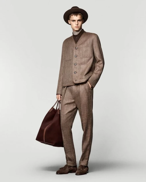 Fashionable adult male in a tailored brown tweed suit with matching hat and brown leather duffel bag, standing in a studio setting.