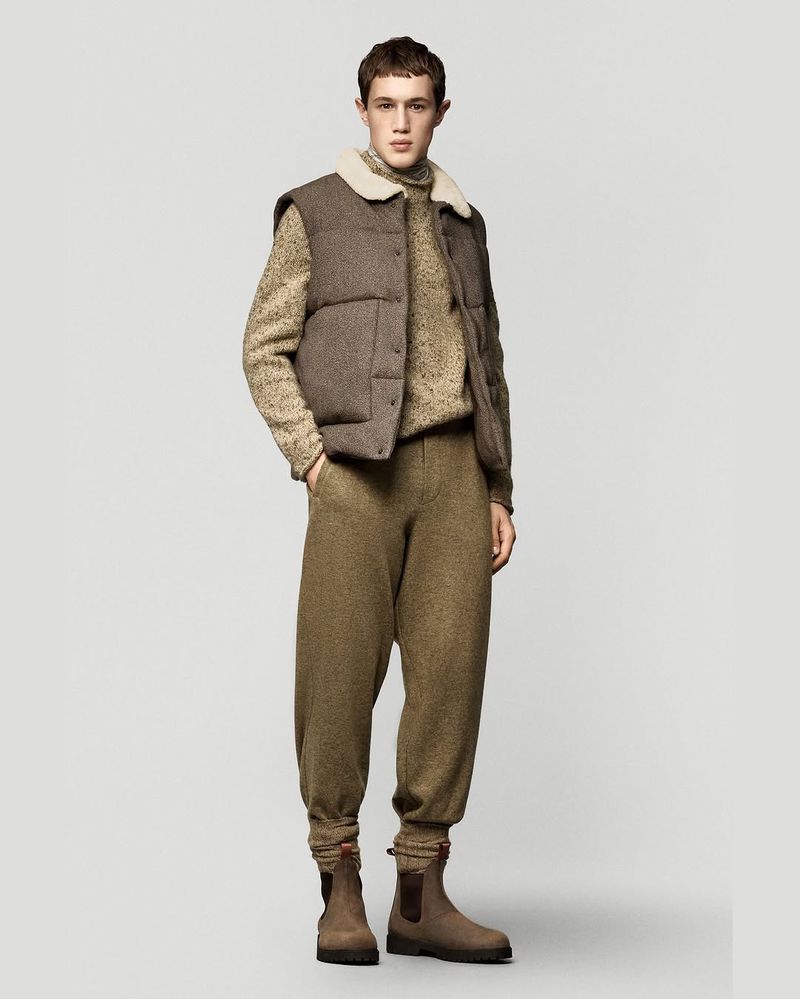 A young man in a puffer vest, sweater, and sweatpants poses against a neutral background, showcasing a blend of casual and comfortable fashion.
