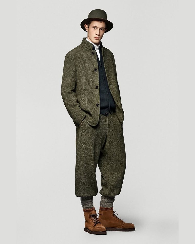 Full-length studio shot of a male model in a green tweed suit, brown boots, and a matching hat, posing against a neutral background.