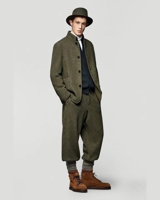 Full-length studio shot of a male model in a green tweed suit, brown boots, and a matching hat, posing against a neutral background.