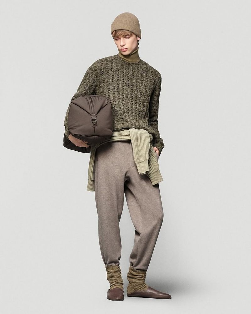 A single adult male poses in a studio setting, wearing a beige beanie, an olive sweater, and brown pants while holding a duffel bag.