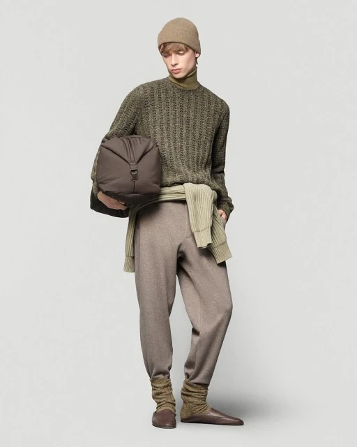 A single adult male poses in a studio setting, wearing a beige beanie, an olive sweater, and brown pants while holding a duffel bag.