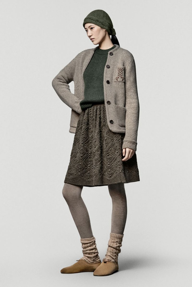 Elegant woman in stylish knitwear: green sweater, patterned skirt, neutral jacket, beanie, and shoes against a plain background.