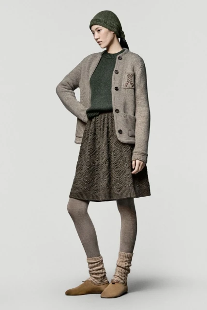 Elegant woman in stylish knitwear: green sweater, patterned skirt, neutral jacket, beanie, and shoes against a plain background.