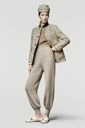Model wearing patterned jacket, matching pillbox hat, and textured knit trousers against a grey seamless background