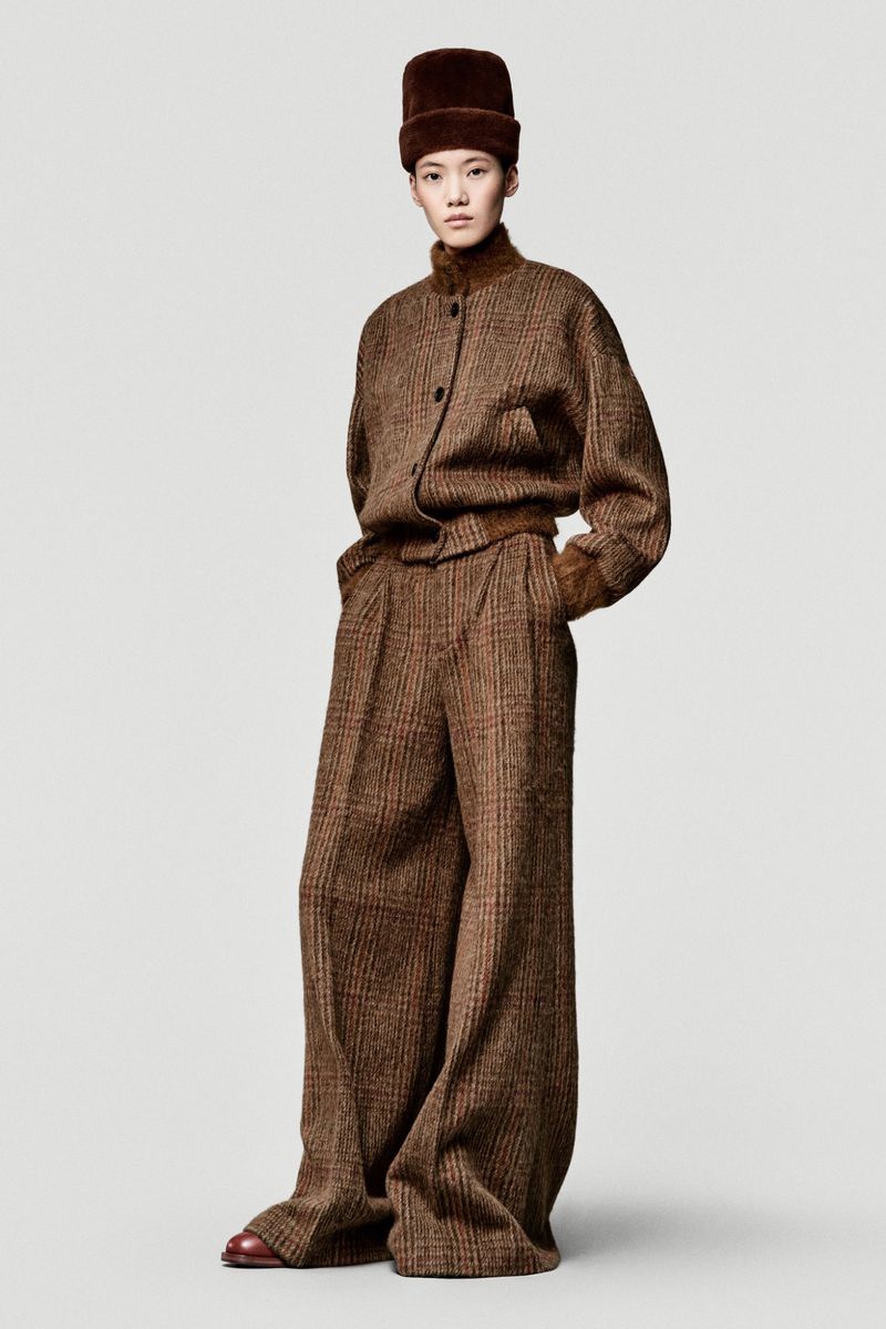 Adult woman poses in wide-legged, plaid tweed pants and matching jacket with fur-trimmed hat in a fashion studio setting.