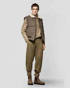 Model wearing a textured brown vest, knit sweater, trousers, and boots against a plain grey seamless background.