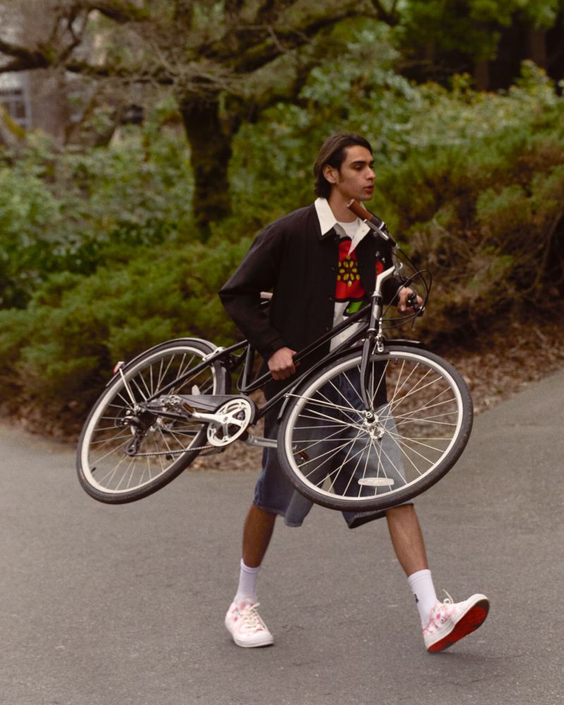 An adult male walks on a paved path carrying a bicycle with green trees in the background during daytime, wearing casual clothing.
